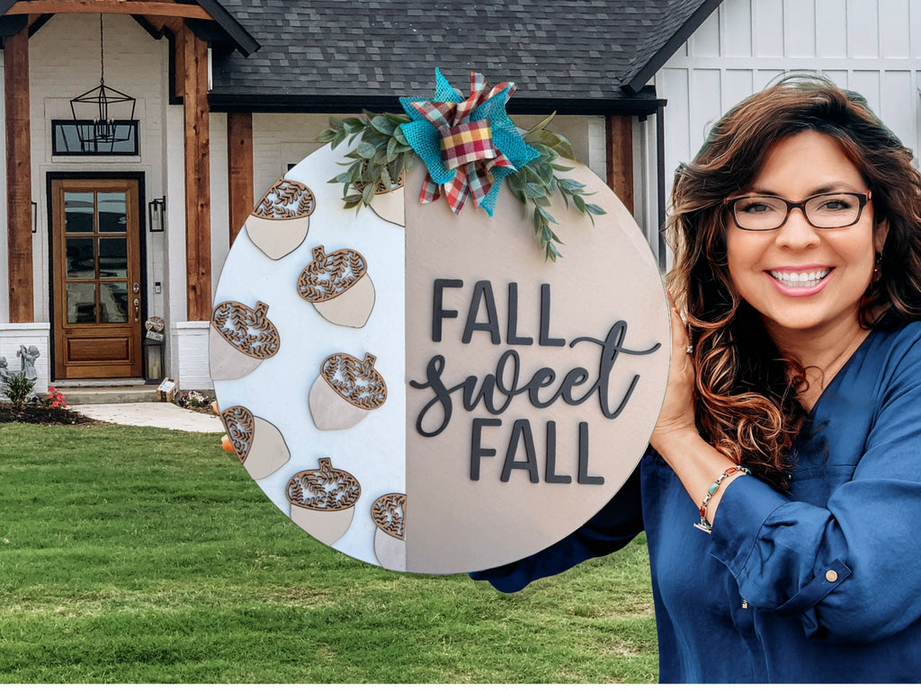 A smiling woman with wavy brown hair and glasses holds a round FALL sweet FALL sign decorated with acorns, leaves, and a plaid bow, standing in front of a modern house with a wooden entryway.
