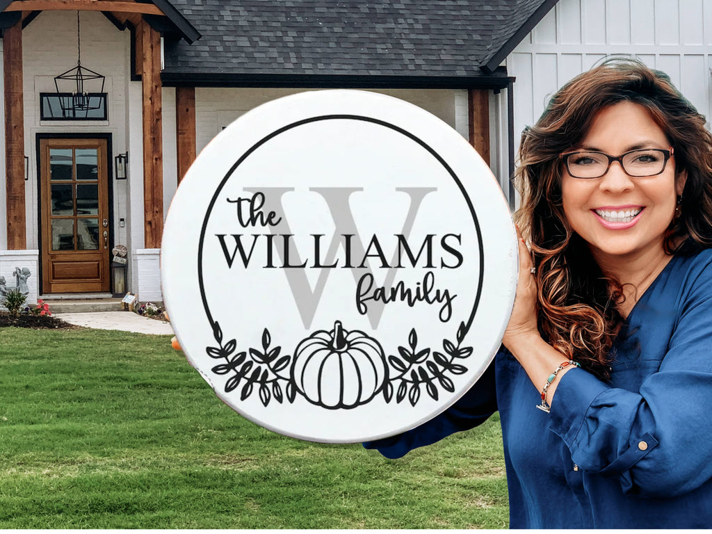 A smiling woman with wavy brown hair and glasses holds a round white sign reading The Williams Family with a large W, leafy accents, and a pumpkin. A modern house with wooden beams and a lawn is in the background.