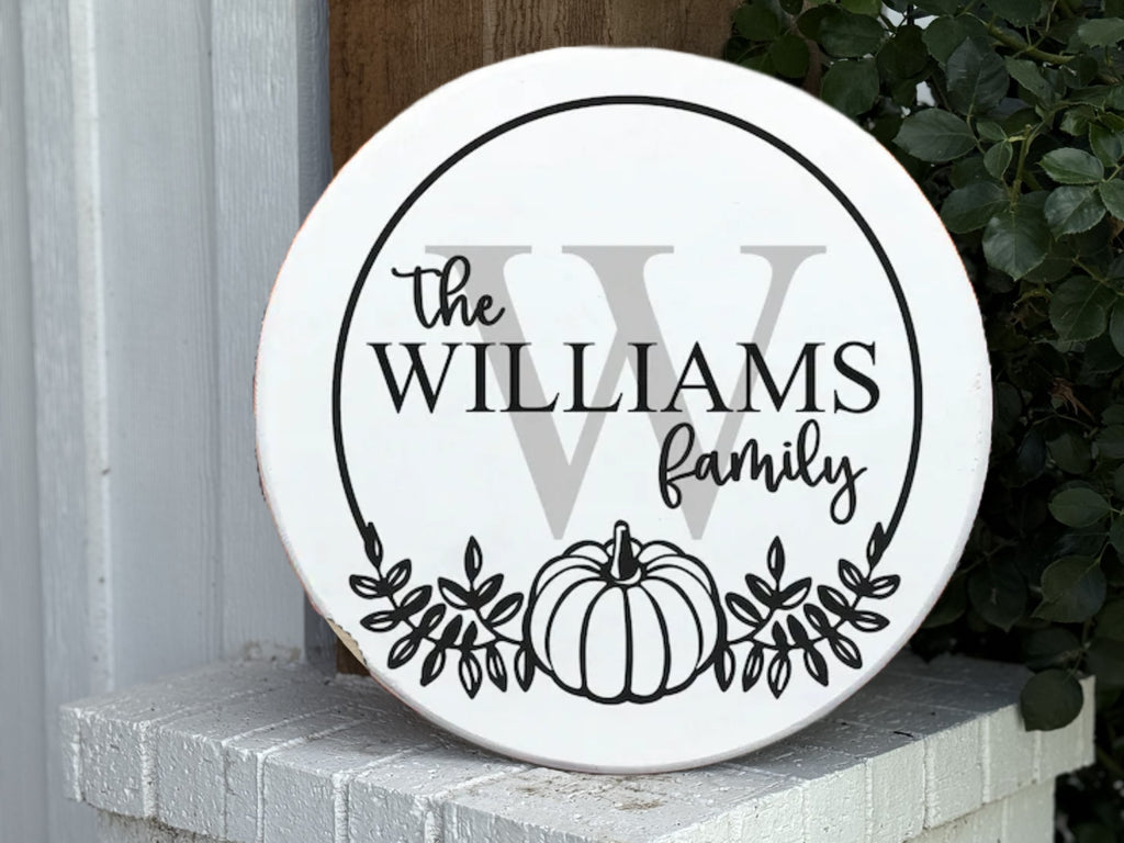 A round white sign with black text that reads The Williams Family over a large gray W, decorated with a pumpkin and leaves at the bottom. The sign rests on a white brick surface near a plant.