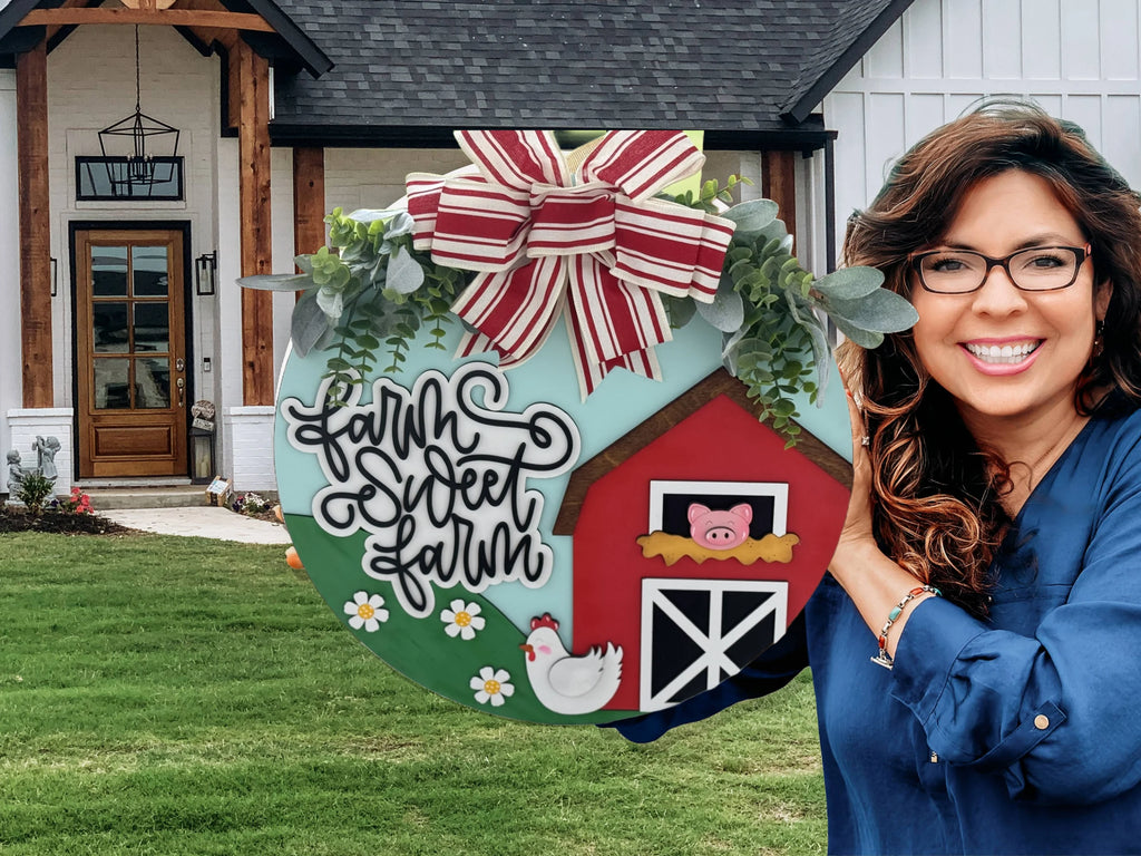 Woman in a blue blouse holding a round farmhouse-themed sign. The sign features a red barn, white fence, pig, hen, daisies, and cursive text reading Farm Sweet Farm. A large striped bow and greenery decorate the top. A farmhouse with wooden and brick accents is in the background.