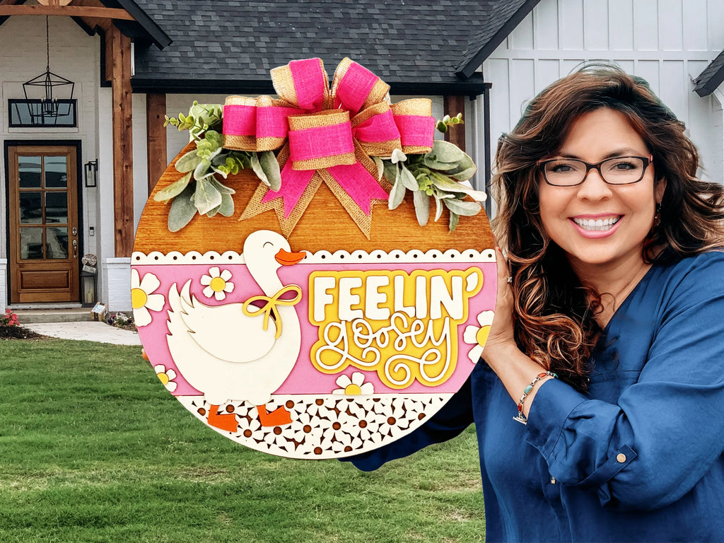 A smiling woman with glasses holds a decorative round sign featuring a duck, flowers, and the words Feelin Goosey, topped with a large pink and gold bow. A modern house is visible in the background.