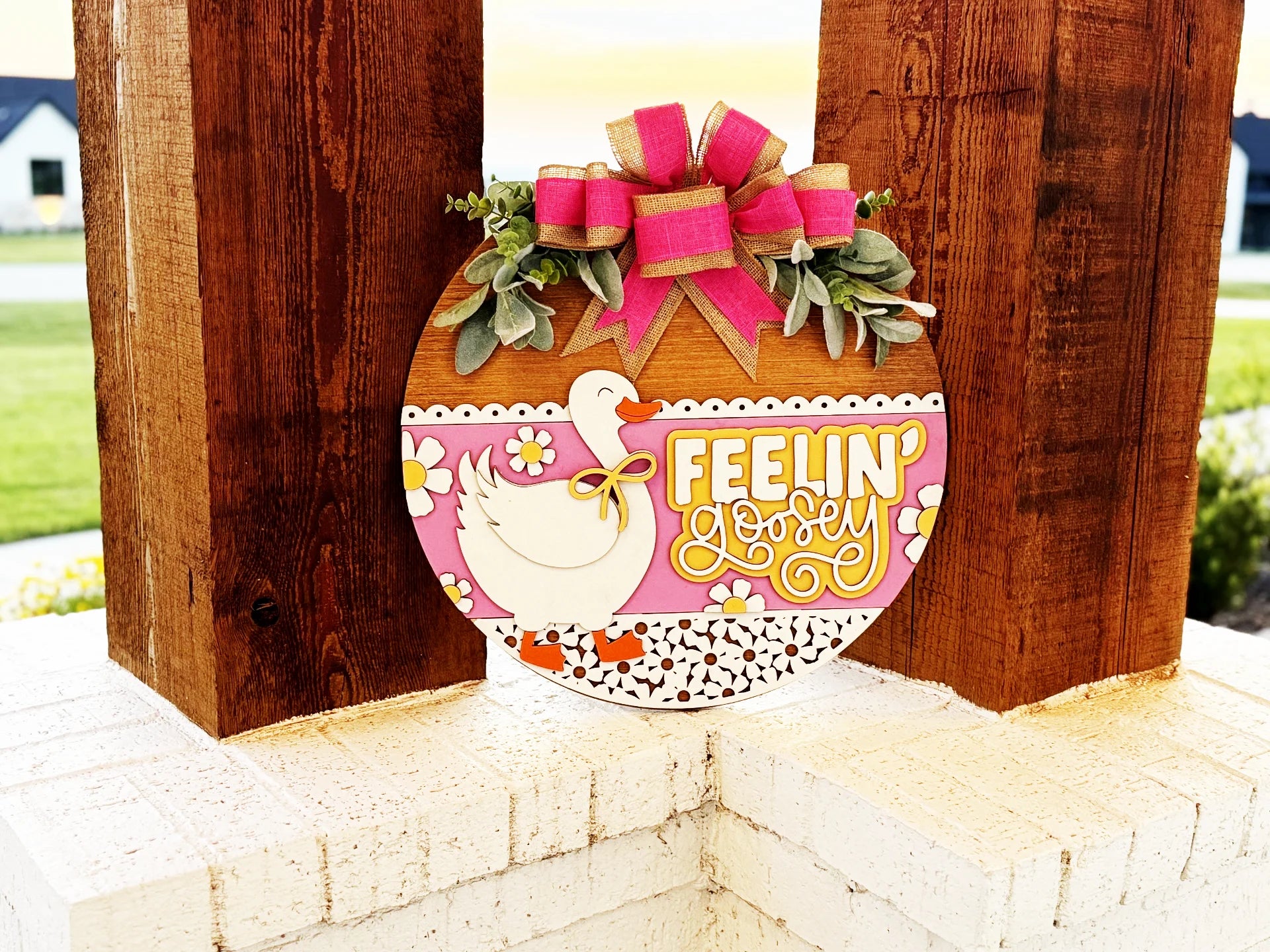 A round wooden sign on a white brick ledge features a duck, pink and yellow flowers, greenery, a pink bow, and the words Feelin Lovely in yellow, set against a sunny outdoor background.