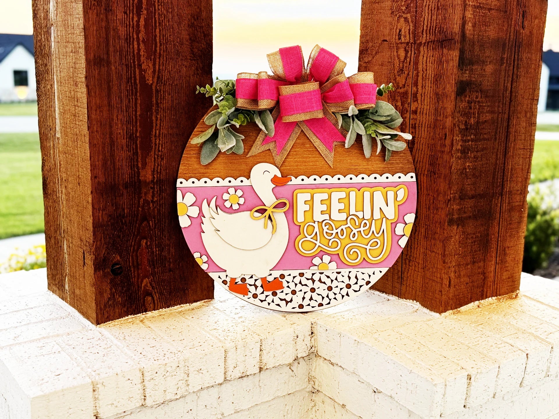 A round wooden sign with a white duck, pink and yellow flowers, and the text Feelin Lovely in yellow, decorated with greenery and a pink bow, hangs on a wooden post outside.