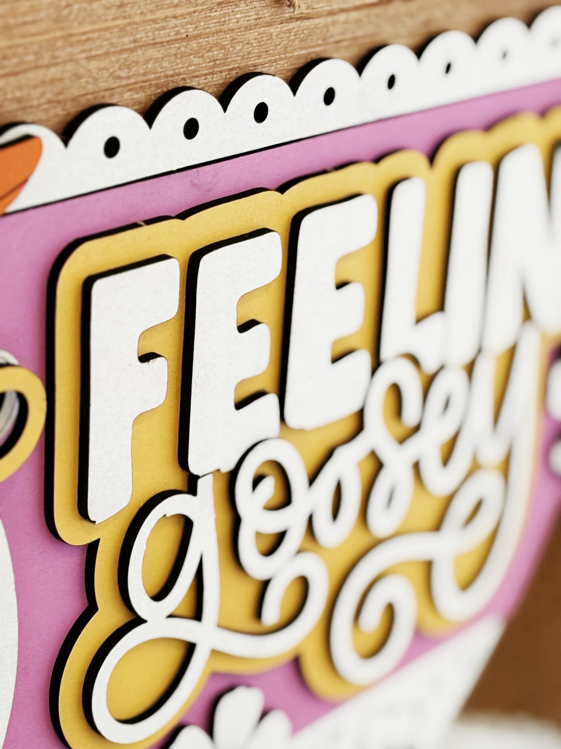 Close-up of a colorful sign with layered, decorative text that reads FEELING goosEY in bold white and yellow letters, with a scalloped white edge and a bright pink background.