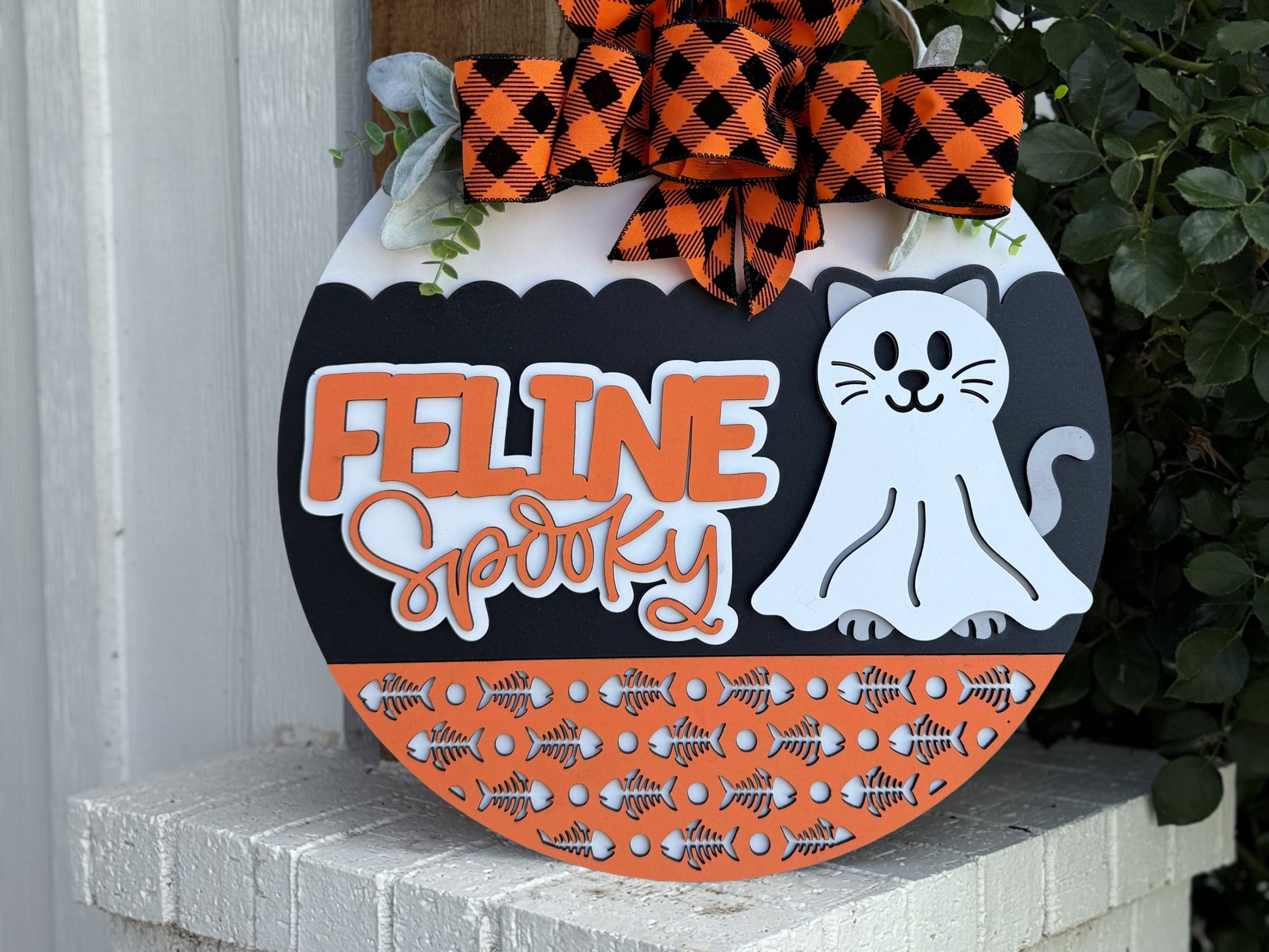 A round decorative sign with an orange and black plaid bow features the words Feline Spooky and an illustration of a white cat. The bottom has a pattern of orange and black fish bones. The sign rests on a white brick surface.