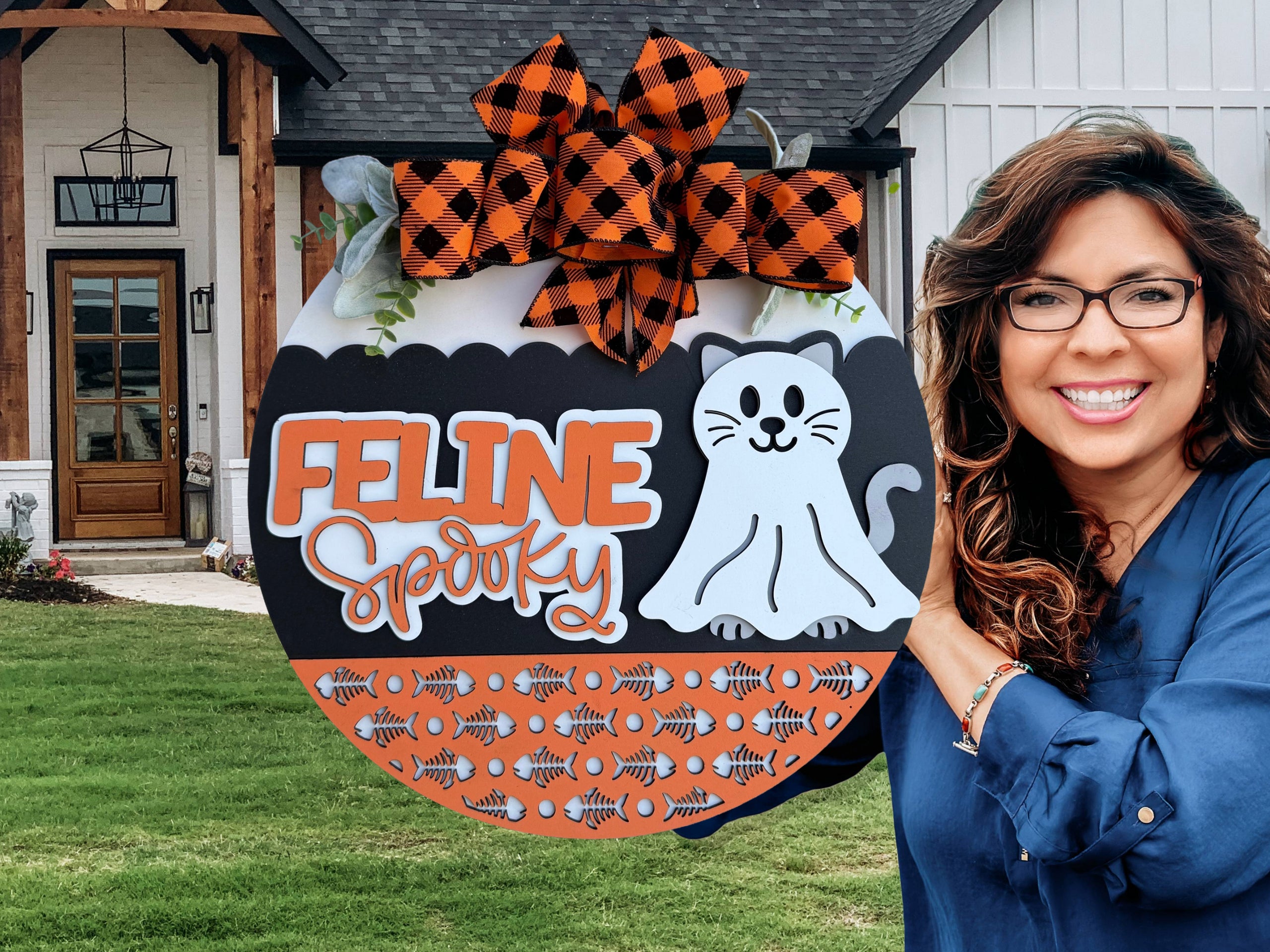 A smiling woman with glasses holds a round Halloween sign decorated with a cartoon cat, orange and black bow, fishbone patterns, and the words FELINE Spooky in front of a modern house.