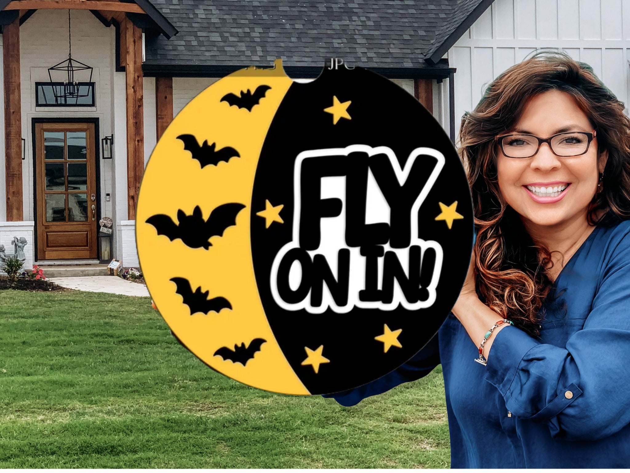 A smiling woman in glasses holds a round Halloween sign with black bats and the words FLY ON IN! in front of a modern house with a green lawn.