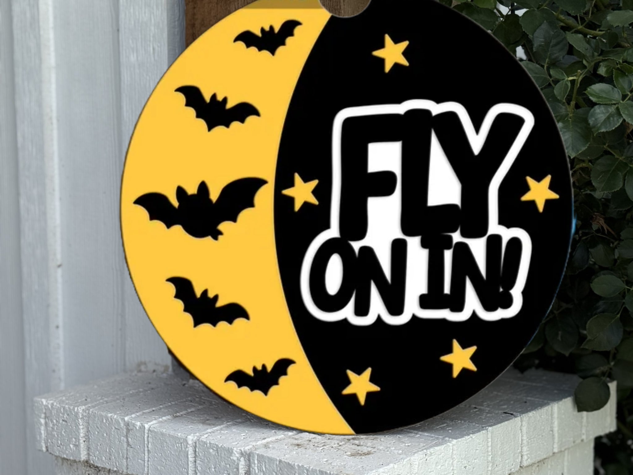 A round sign with a yellow and black design features black bats, yellow stars, and bold white text reading “FLY ON IN!”. The sign leans against a white brick surface near green foliage.