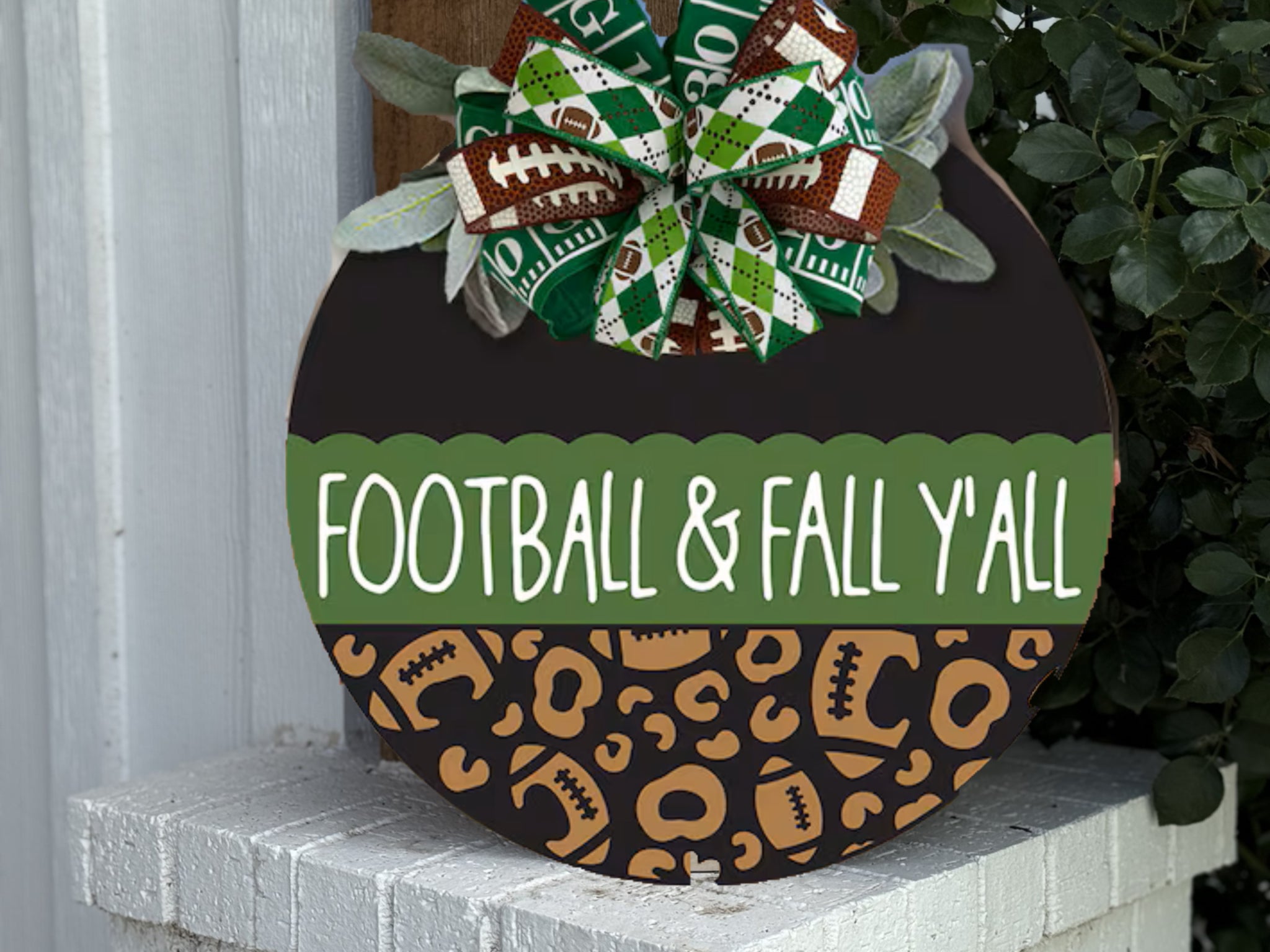 A round wooden sign decorated with a green and brown bow on top. The sign reads “FOOTBALL & FALL Y’ALL” and features football and leopard print designs on the bottom half. It rests on a white brick ledge.