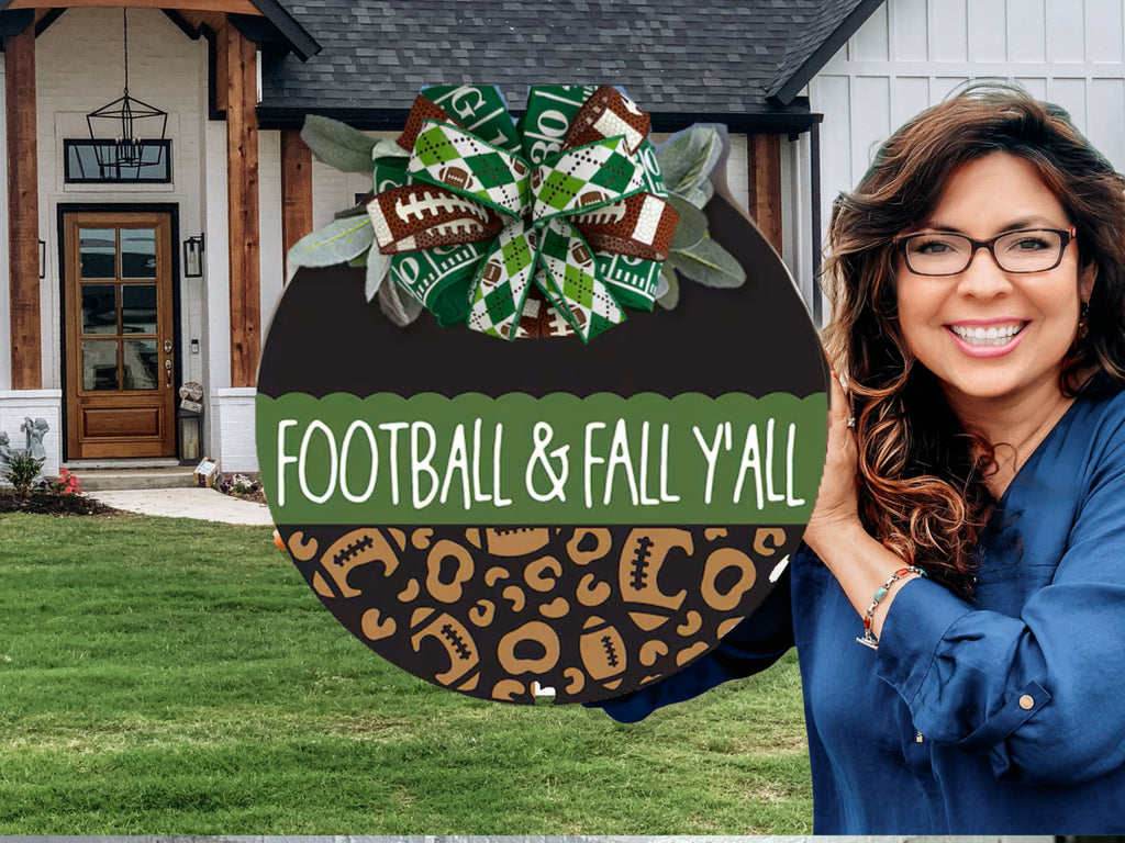 A smiling woman with long wavy hair and glasses holds a round sign decorated with football and leopard patterns, reading “FOOTBALL & FALL Y’ALL,” in front of a modern house with wooden and white accents.