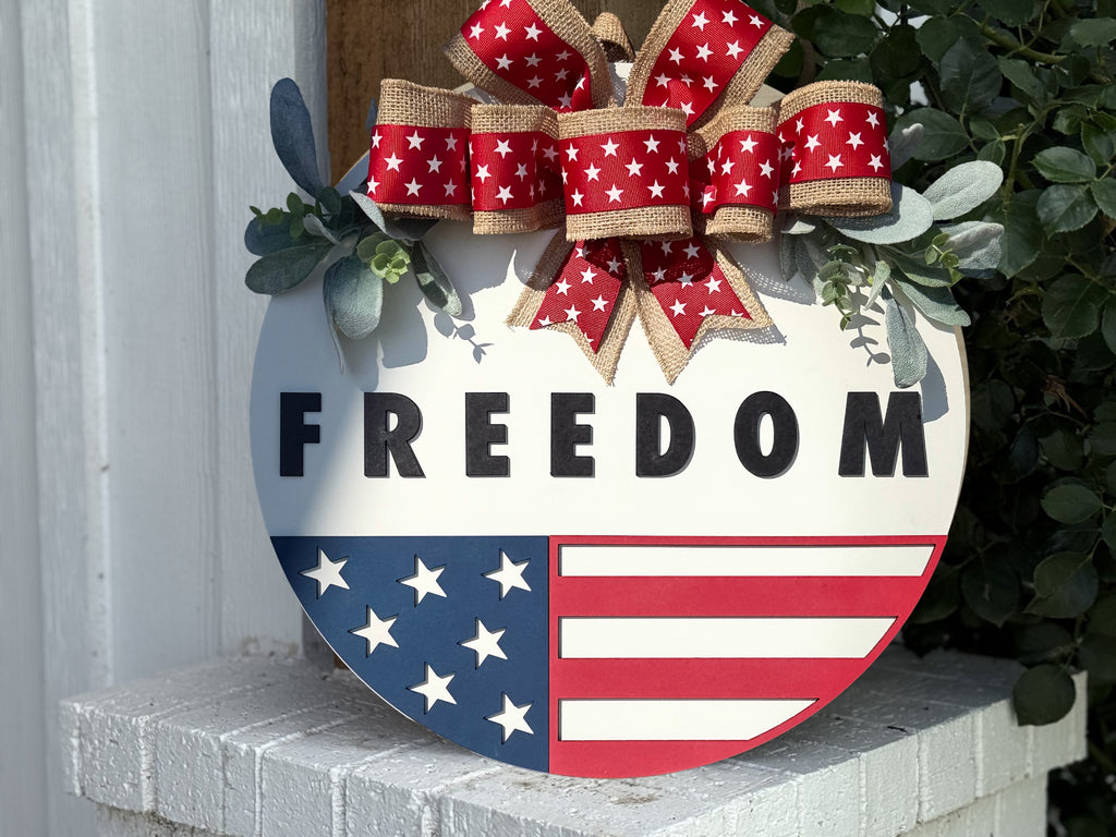 A round sign with FREEDOM written in bold black letters, decorated with an American flag design on the bottom half and a red, white, and burlap bow with stars on top, placed on a white brick surface.
