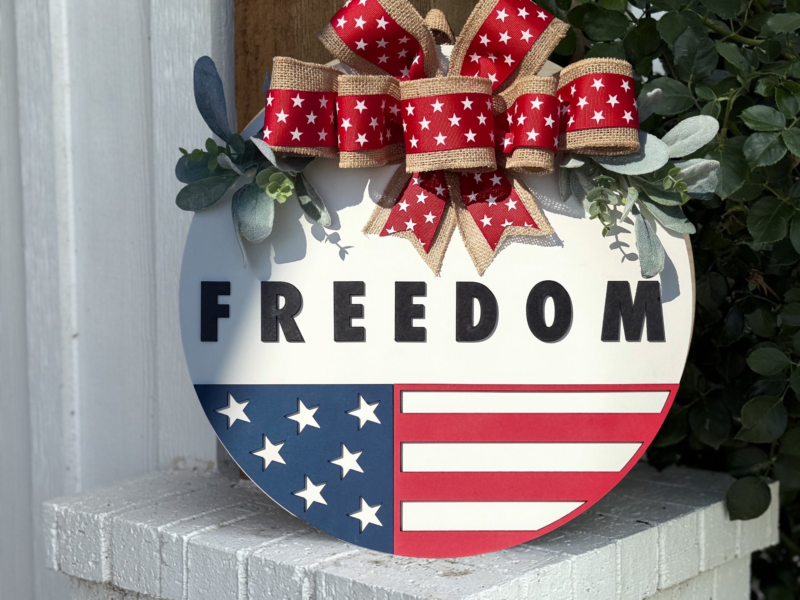 A round sign with FREEDOM written in bold black letters, decorated with an American flag design on the bottom half and a red, white, and burlap bow with stars on top, placed on a white brick surface.