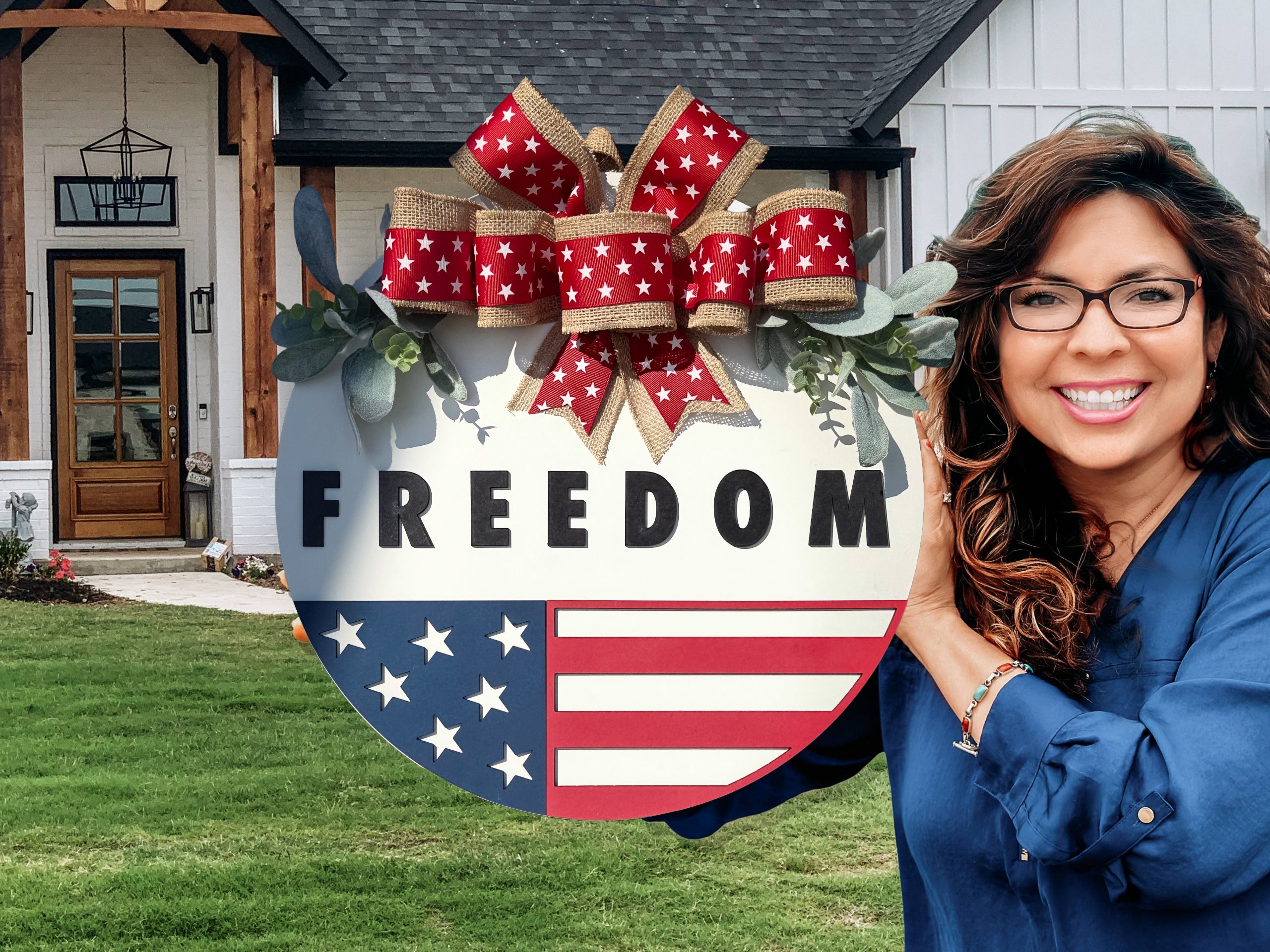 A smiling woman in glasses holds a round sign decorated with stars, stripes, and a red, white, and blue bow. The sign reads FREEDOM. She stands in front of a modern house with a green lawn.