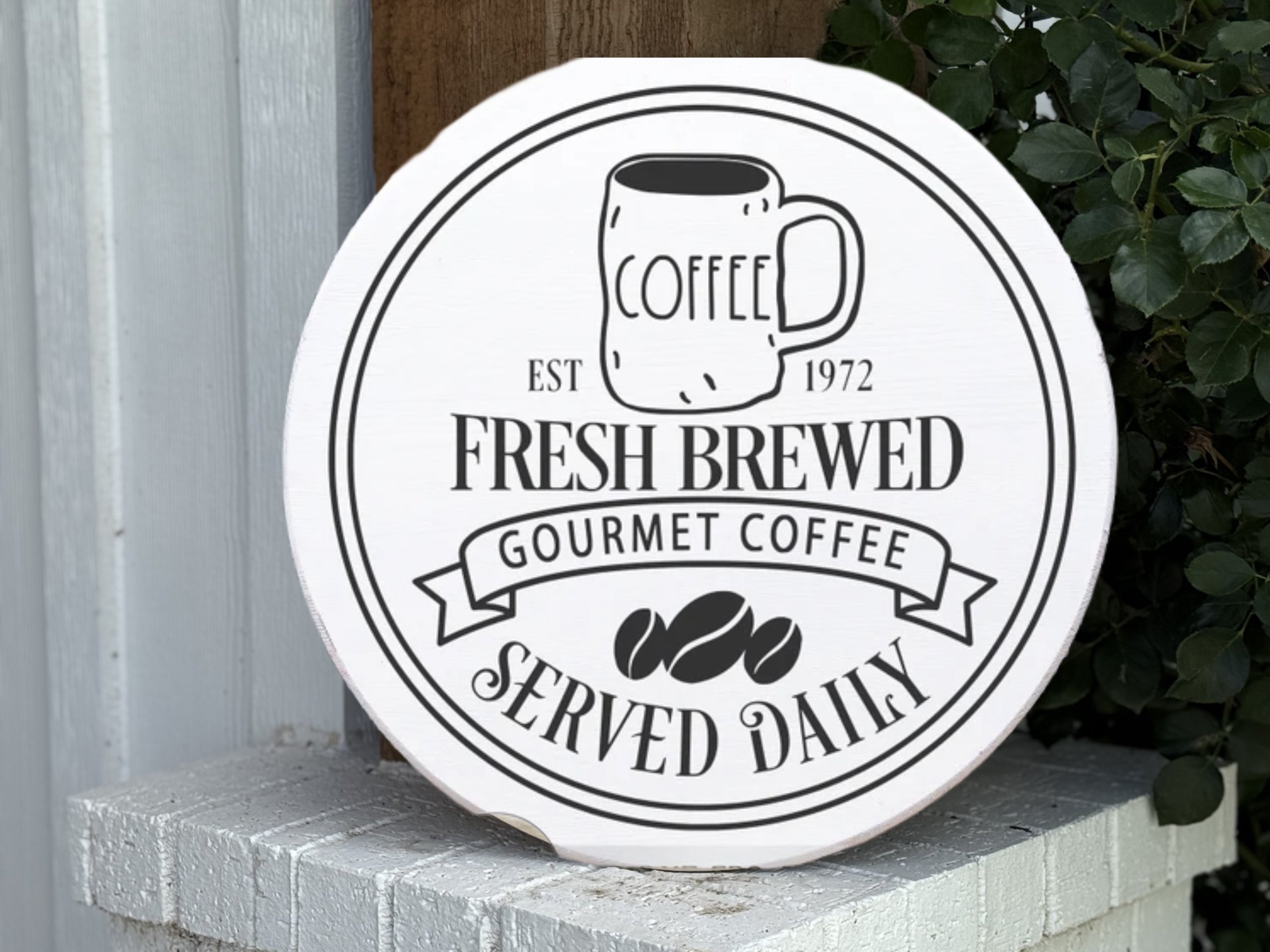 A round white sign with a coffee mug illustration and text reading: COFFEE EST 1972 FRESH BREWED GOURMET COFFEE SERVED DAILY, displayed on a white brick ledge beside green leaves.