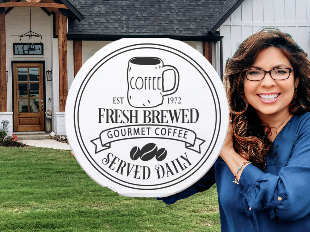 A smiling woman with long brown hair and glasses holds a round sign that reads Fresh Brewed Gourmet Coffee Served Daily in front of a modern house with a wooden porch.