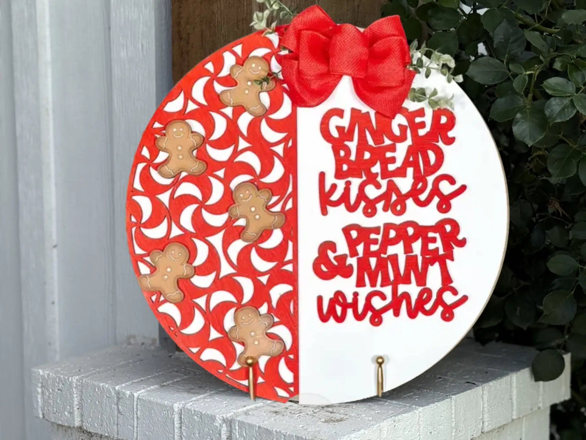 A round holiday sign with a red bow, half decorated with red and white peppermint swirl patterns and gingerbread men, and the other half with the words “Gingerbread kisses & peppermint wishes” in red letters.