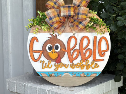 A round wooden sign with a cartoon turkey face, autumn plaid bow, and the words “Gobble ’til you wobble” in orange and yellow letters, hangs on a white porch with green leaves in the background.