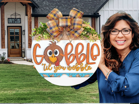A smiling woman stands outside a house, holding a round Thanksgiving sign decorated with a cartoon turkey, greenery, a plaid bow, and the words Gobble til you wobble.