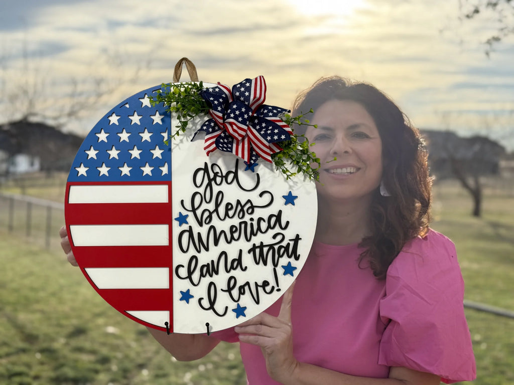 A woman in a pink blouse stands outdoors, holding a circular plaque. The left side resembles an American flag, while the right side has the handwritten text God bless America land that I love! A decorative bow with stars and stripes and greenery adorns the top. The background is a sunny yard.