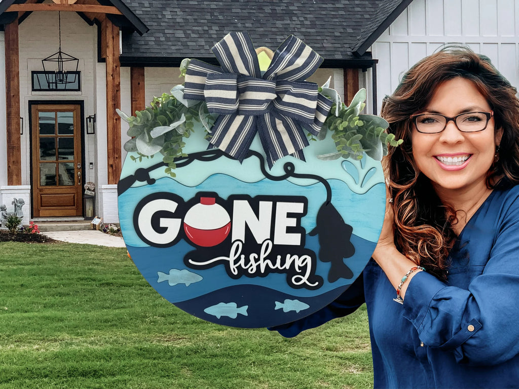 A person in a blue shirt holds a circular turquoise sign with Gone fishing written on it, decorated with fish and a bobber. The sign has a striped bow and foliage. They stand in front of a house with a wooden door and green lawn.
