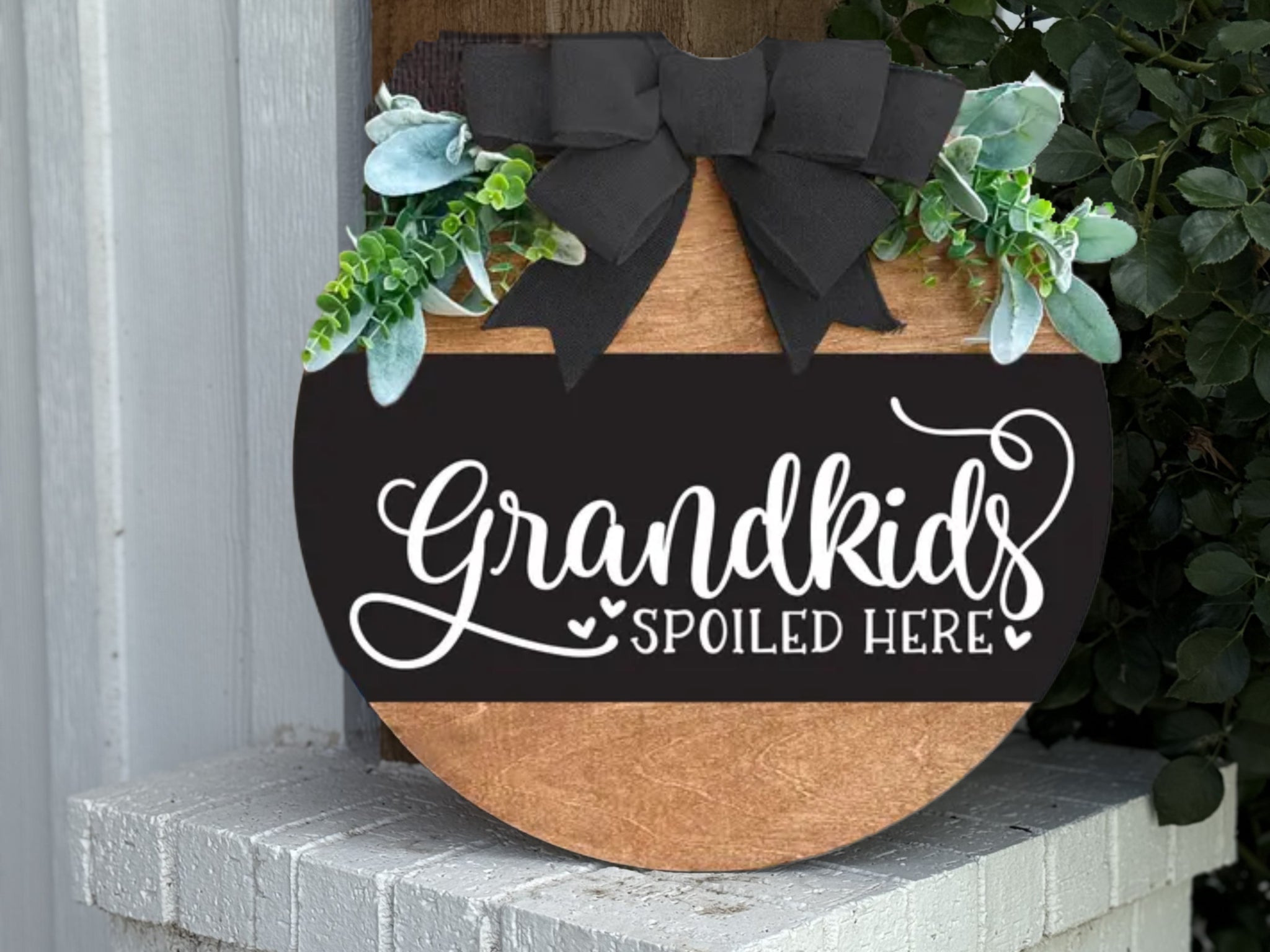 A round wooden sign with greenery, a black ribbon, and white script reads “Grandkids Spoiled Here.” The sign is propped up on a white brick surface next to a vertical wooden post.