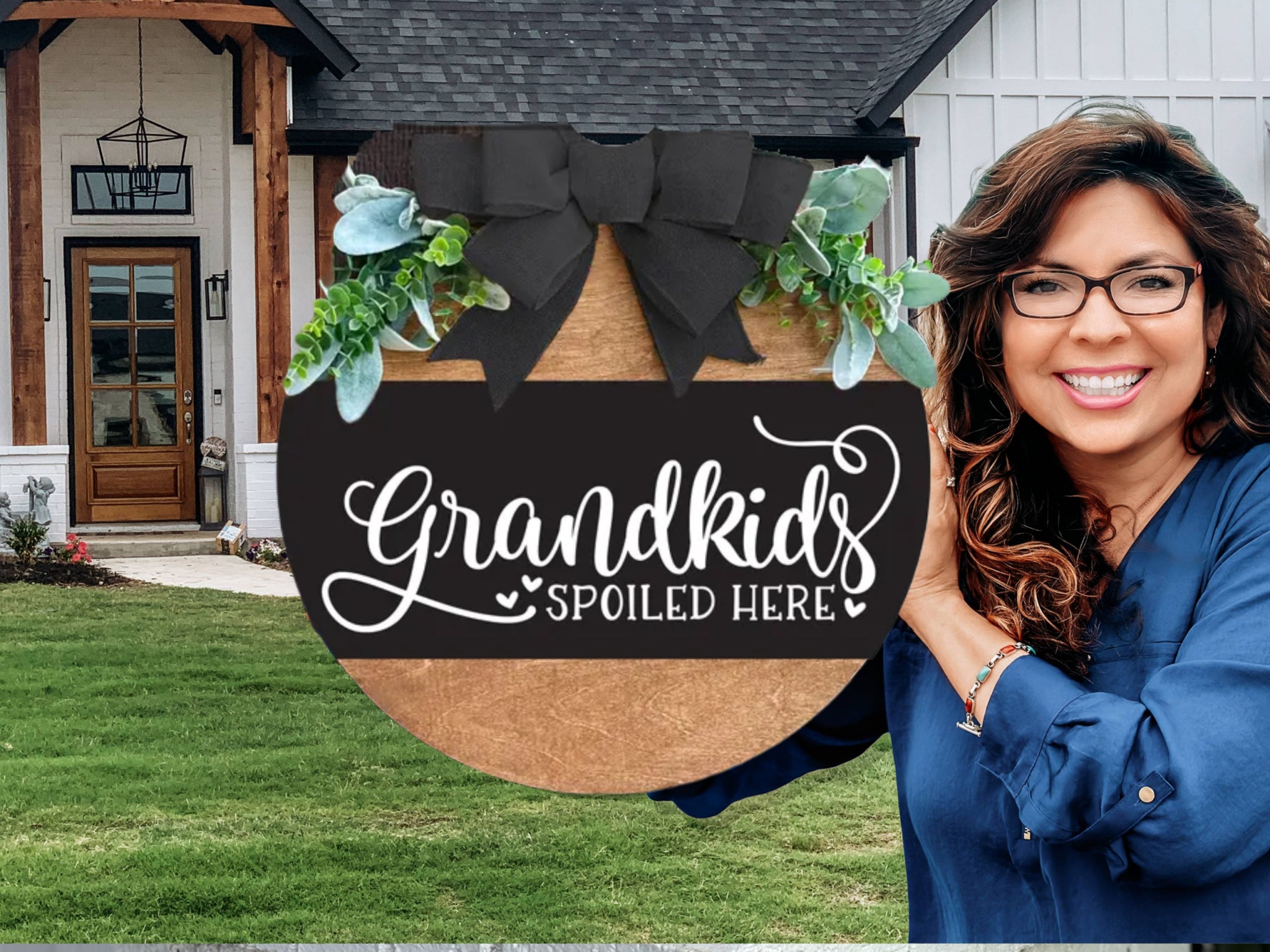 A smiling woman in glasses and a blue shirt holds a round wooden sign with greenery and a black bow, reading “Grandkids Spoiled Here,” standing outside a modern house with a wooden door and green lawn.