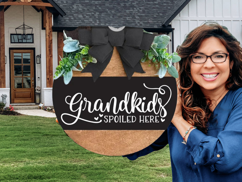 A smiling woman holds a round wooden sign decorated with greenery and a black bow, reading “Grandkids Spoiled Here,” in front of a modern house with a neat lawn and wooden accents.