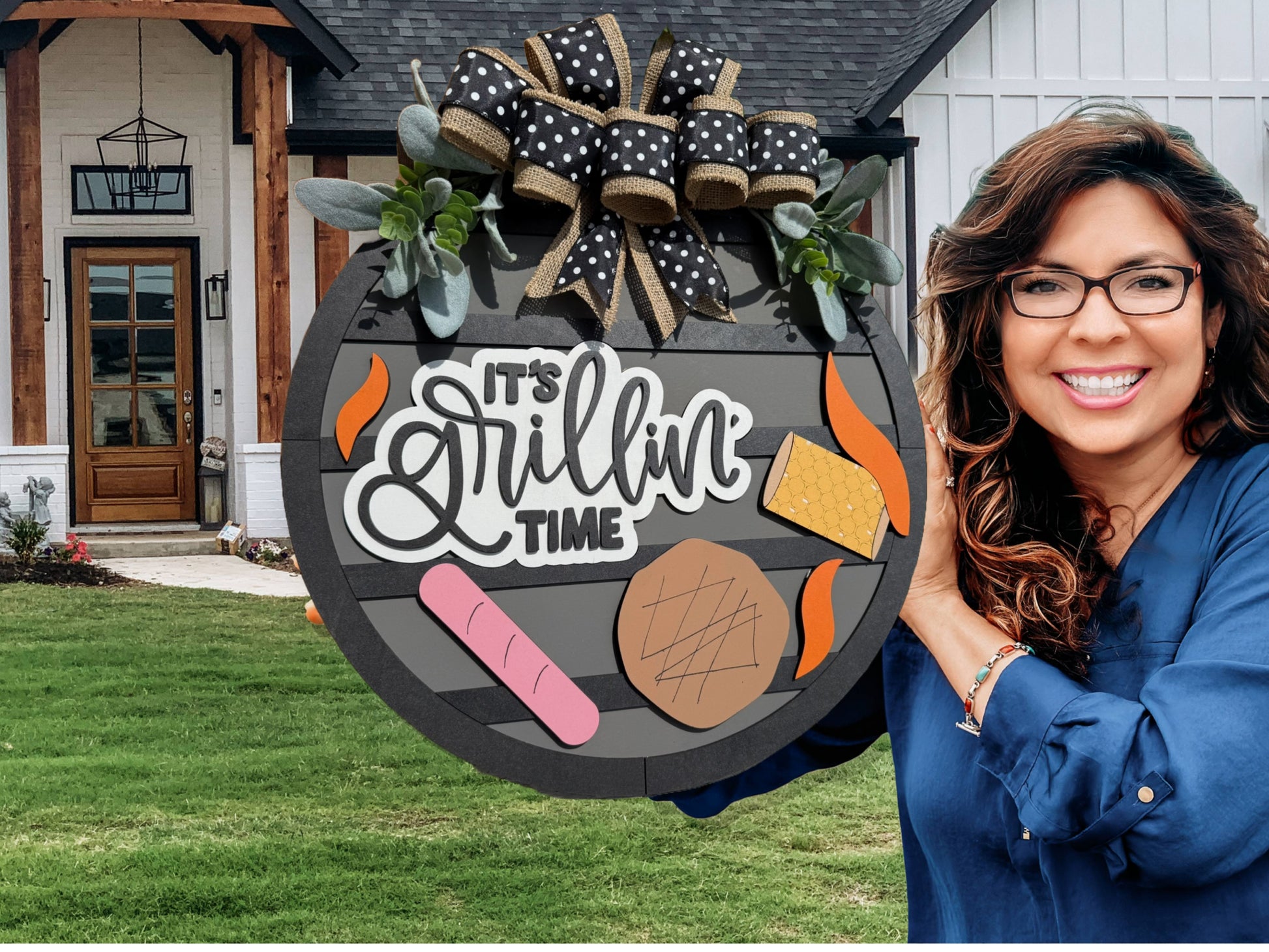 A smiling woman with long brown hair and glasses holds a round sign that says Its Grillin Time with images of grilled food, standing in front of a modern house with a green lawn.