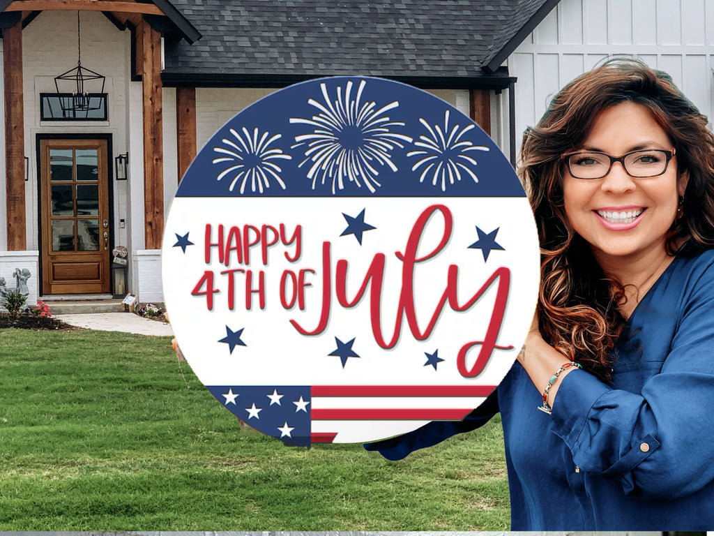 A smiling woman holding a round sign that says Happy 4th of July with fireworks and stars, standing in front of a modern house with a green lawn.