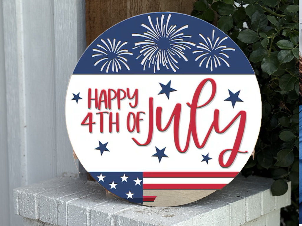 A round sign features fireworks, stars, and stripes with the text Happy 4th of July in red and blue, displayed outdoors on a white brick ledge next to green leaves.