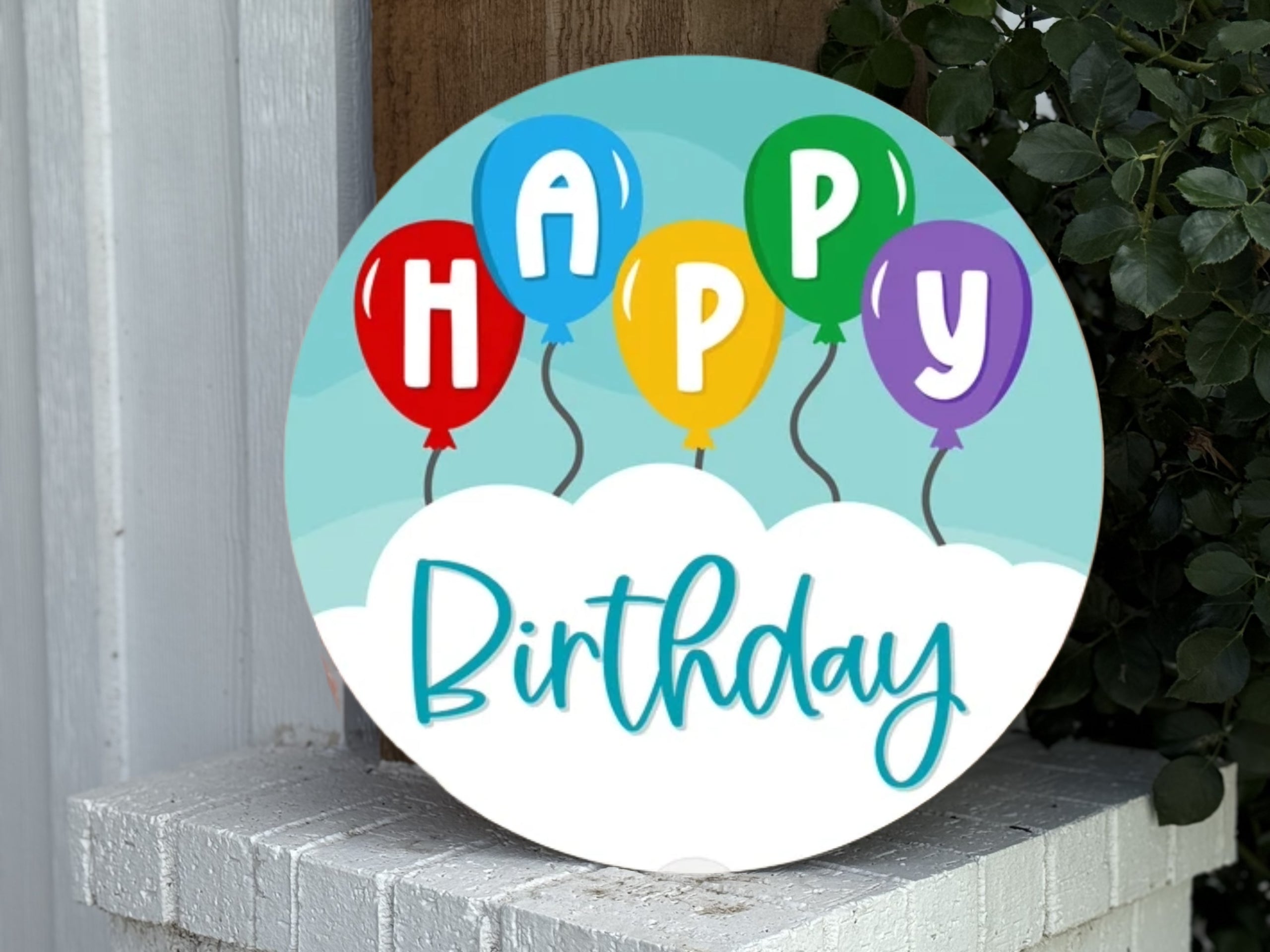 A round sign with colorful balloons spelling “HAPPY” above the word “Birthday” in blue script, set against a sky and cloud background, propped on a white brick surface near greenery.