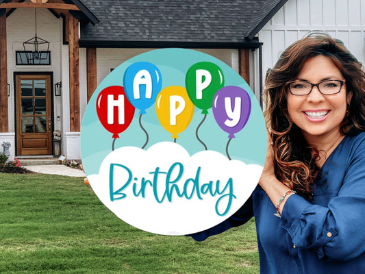 A smiling woman stands outside a house holding a colorful sign with balloons that says Happy Birthday.