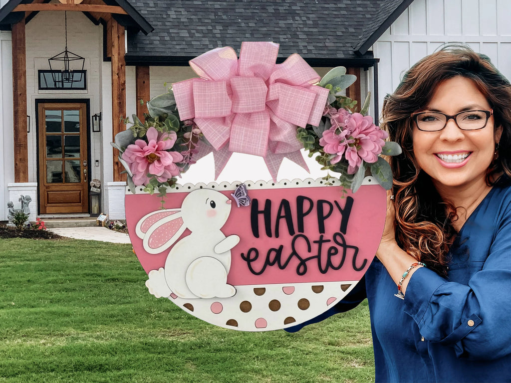 A smiling woman with wavy brown hair and glasses holds a round pink and white Easter wreath decorated with a bunny, pink flowers, a large pink bow, gold polka dots, and the words “Happy Easter,” standing in front of a modern house with white siding.