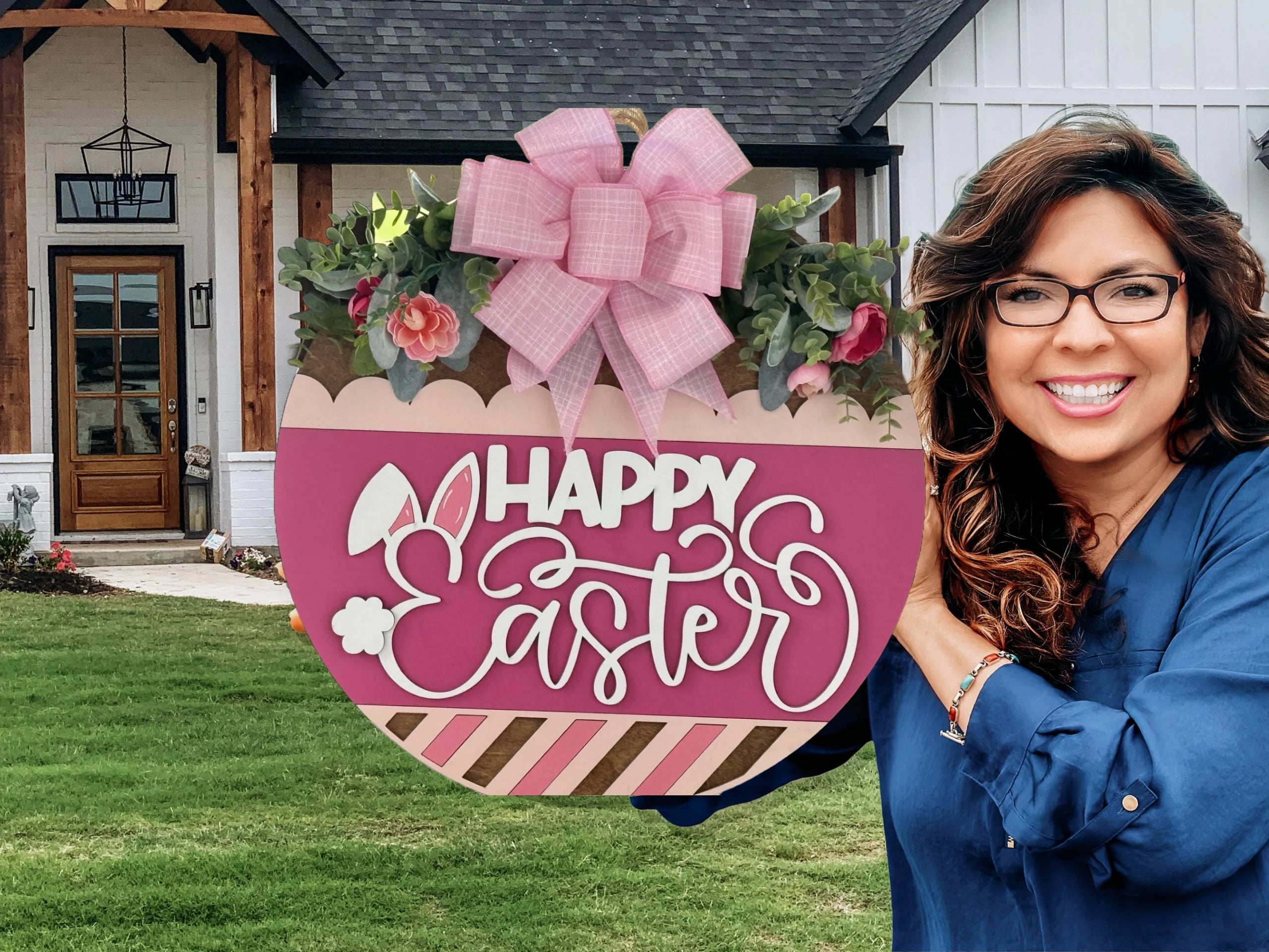 A smiling woman with long brown hair and glasses, wearing a blue shirt, holds a large pink Easter sign shaped like an egg with a pink bow, flowers, and the words Happy Easter. She stands outside a modern house with a wooden door and manicured lawn.