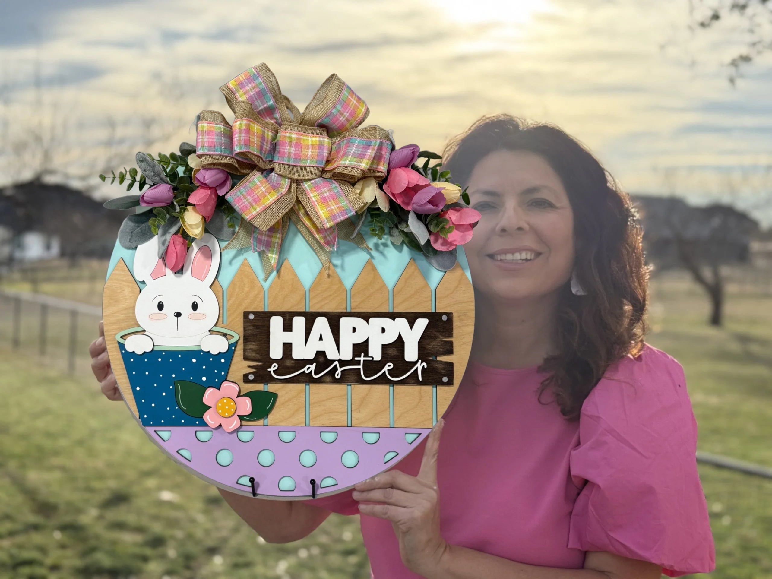 A woman in a pink shirt holds a colorful Easter sign. The sign features a white bunny, flowers, and the words Happy Easter on a wooden fence background. A pink and yellow plaid bow decorates the top. The setting is an outdoor grassy area with a soft-focus background.