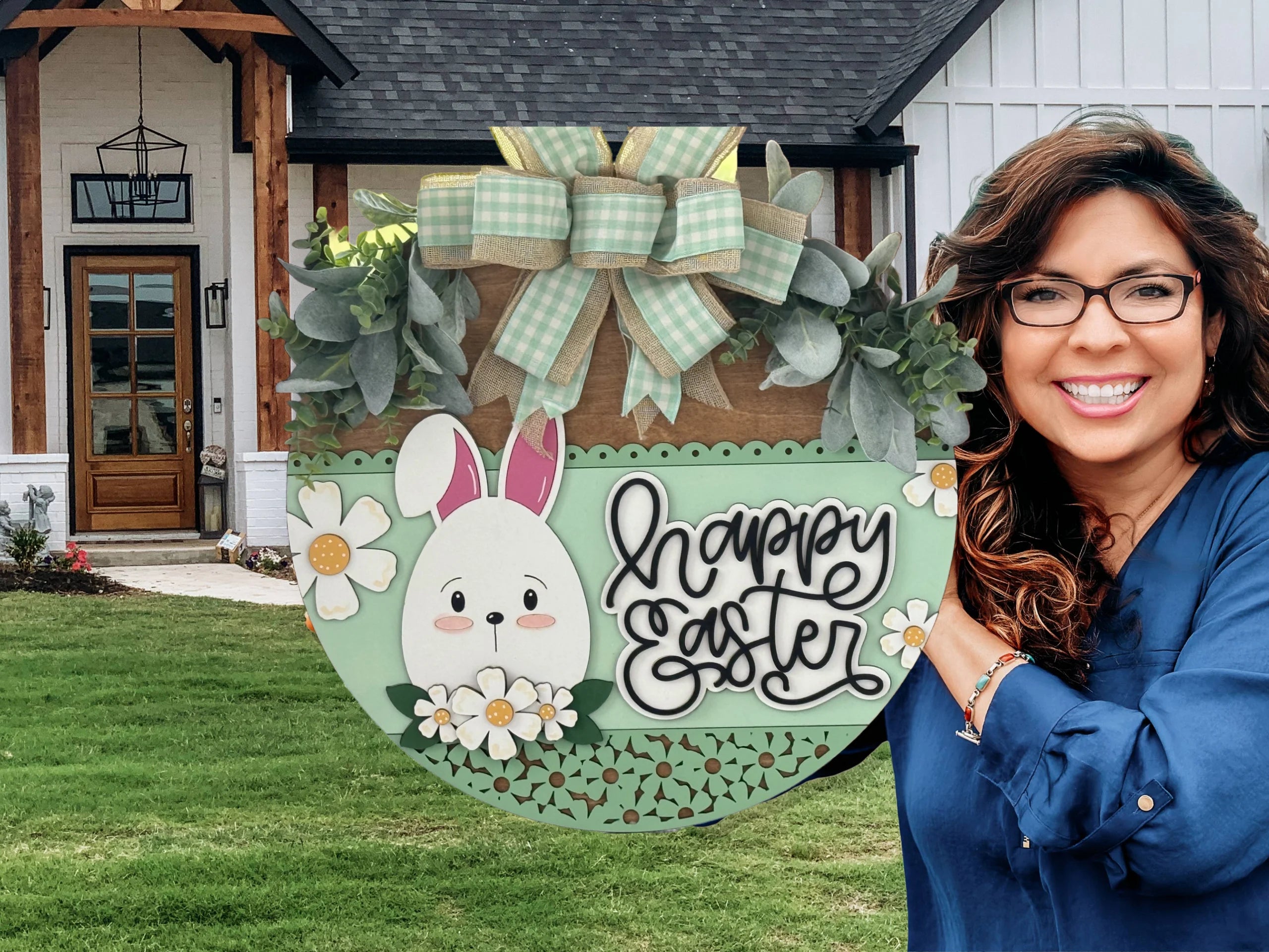 A smiling woman with glasses holds a decorative Happy Easter sign featuring a bunny, flowers, and green bows. She stands on a green lawn in front of a modern farmhouse with wood and white accents and a glass-panel front door.