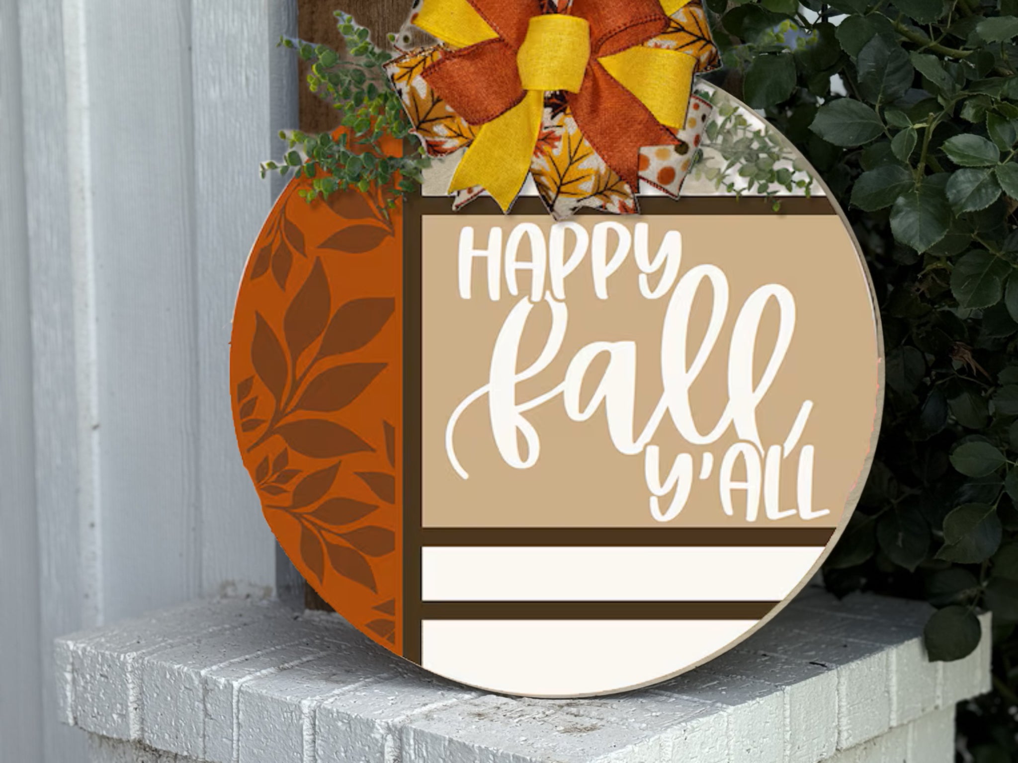 A round decorative sign with a bow and leaves reads Happy fall y’all in white script, featuring autumn colors and leaf designs, displayed on a white outdoor ledge near greenery.