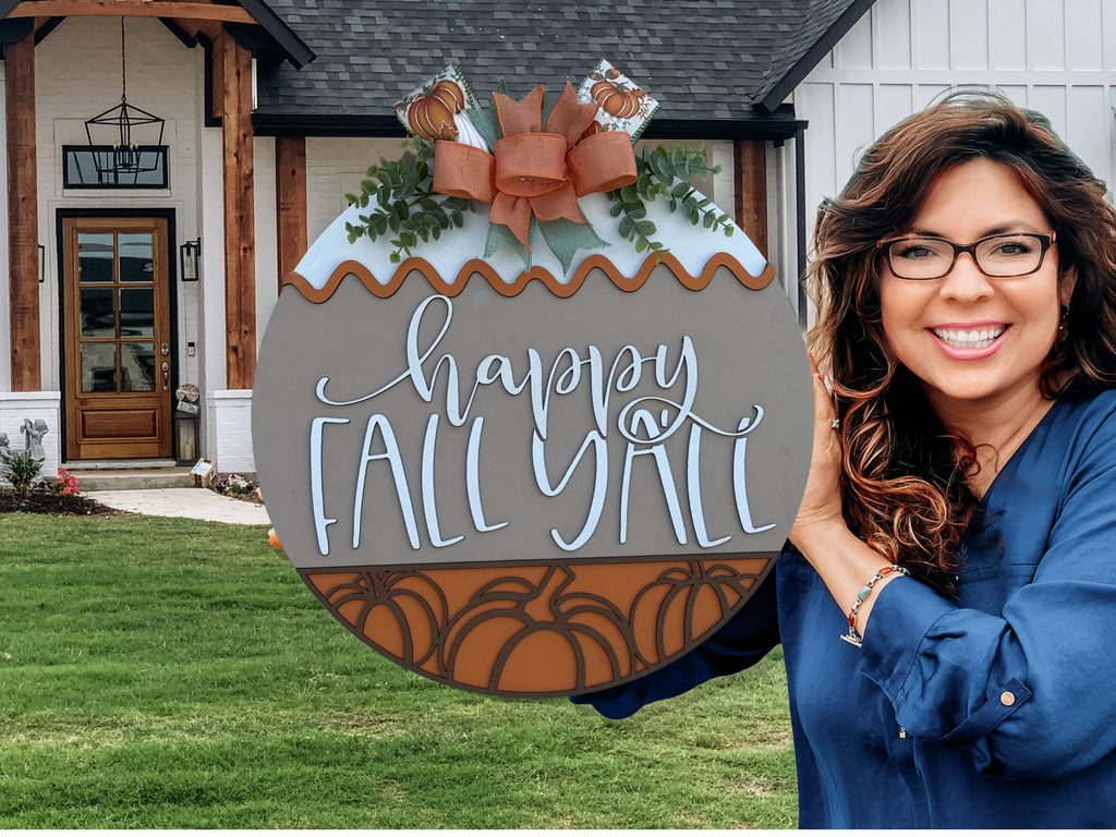 A smiling woman in a blue shirt stands outside a house, holding a large round sign decorated with pumpkins, acorns, and a bow, reading Happy Fall Yall.