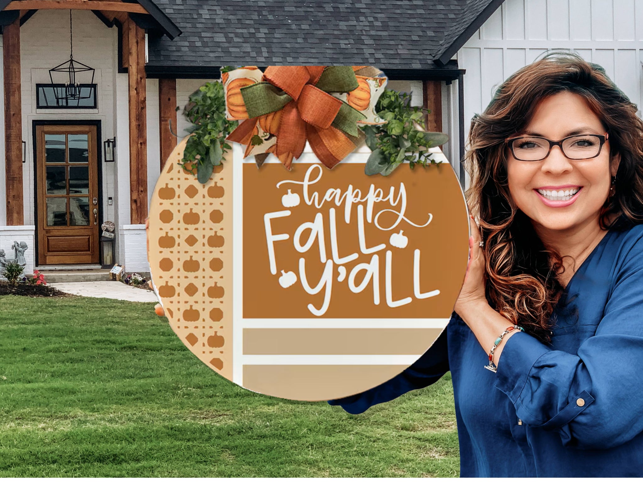 A smiling woman in glasses holds a round sign with pumpkins, greenery, and a bow that reads Happy Fall Yall. She stands in front of a modern house with a green lawn and wooden accents.