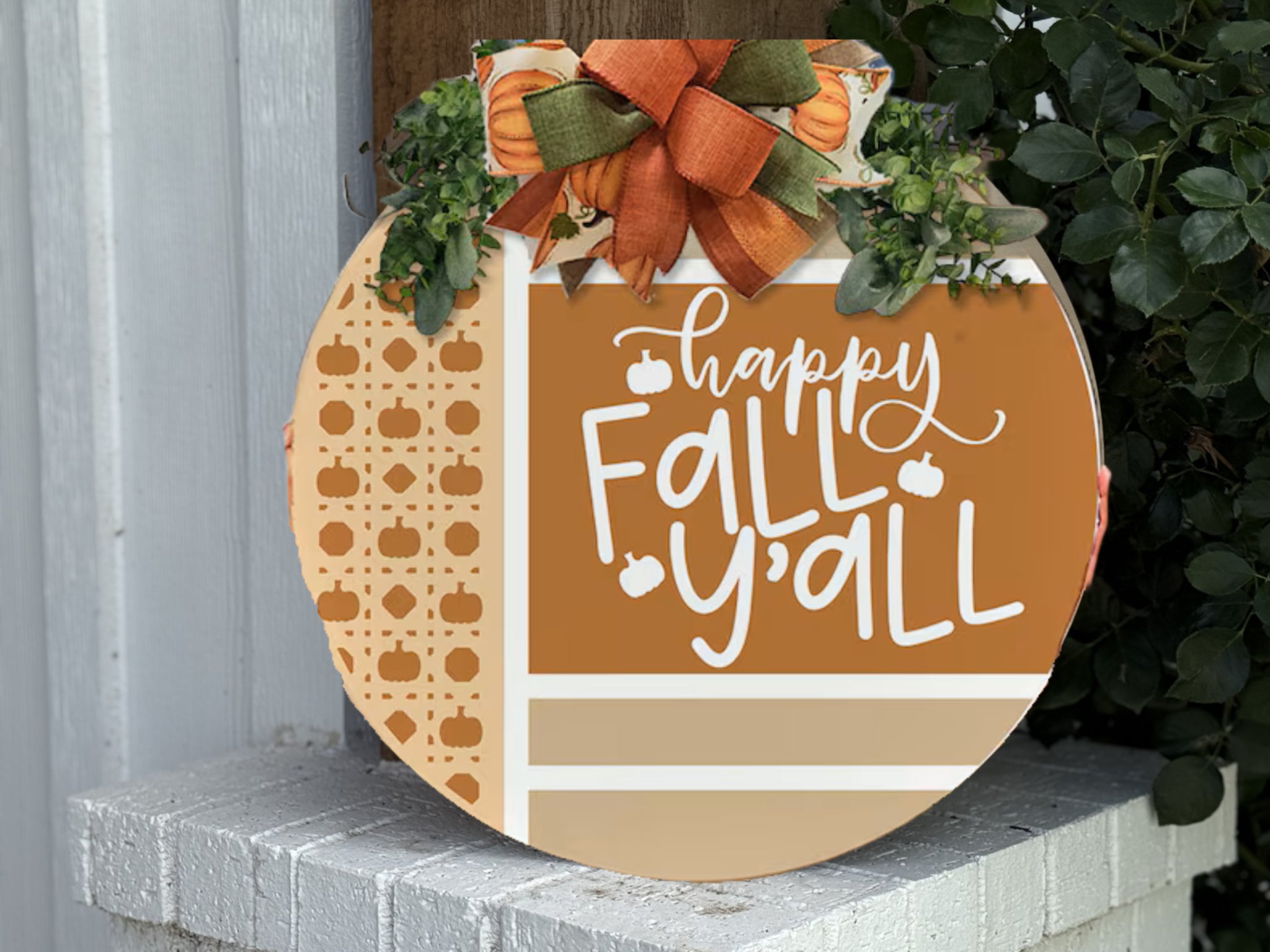 A round autumn-themed sign with a bow and greenery reads “Happy Fall Y’all” in white script on an orange background, decorated with small pumpkin patterns, leaning against a white brick ledge near some plants.