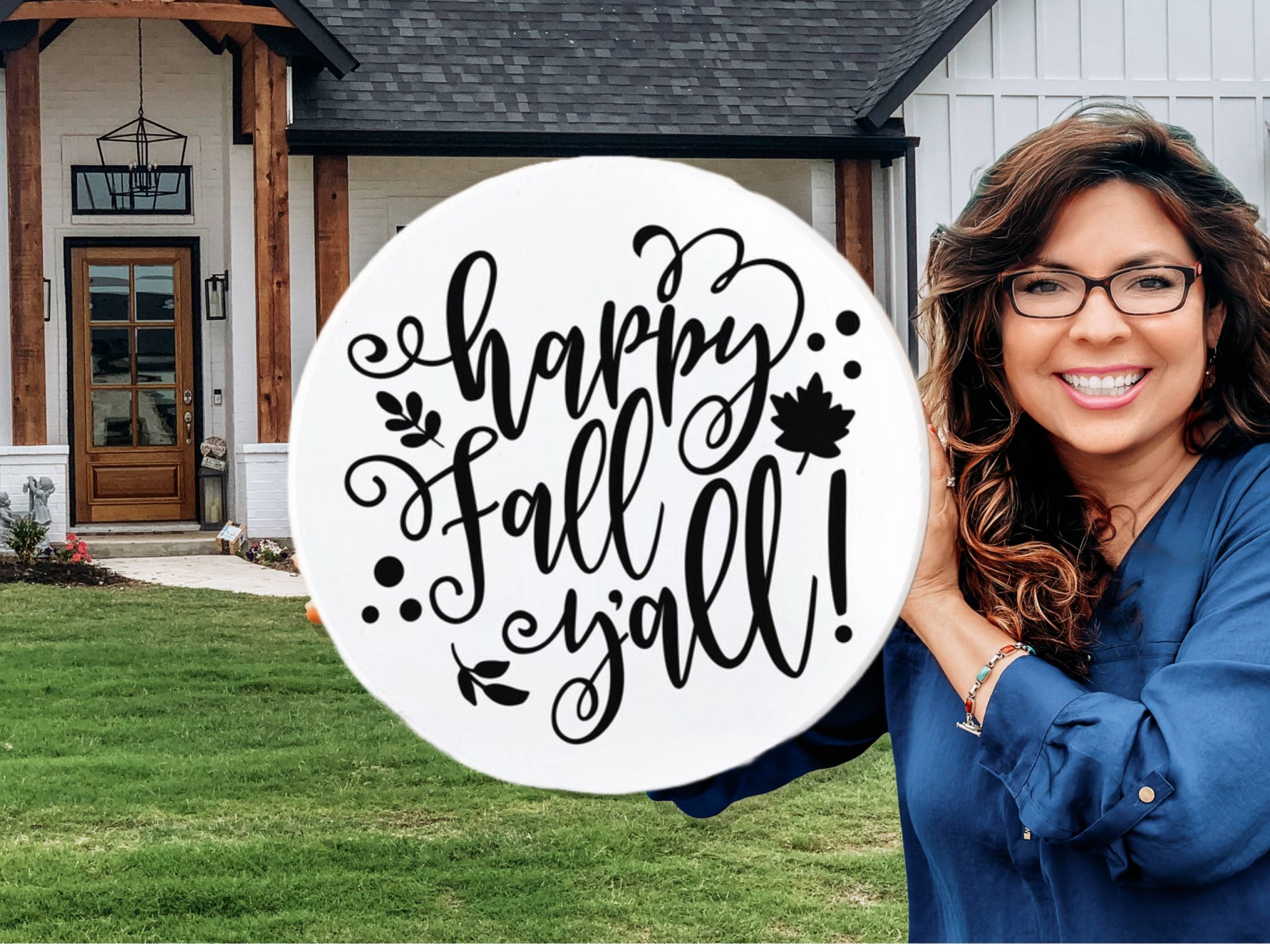 A smiling woman with long wavy hair and glasses holds a round sign reading Happy Fall Yall! in front of a modern house with a lawn. The sign is decorated with leaves and swirls.