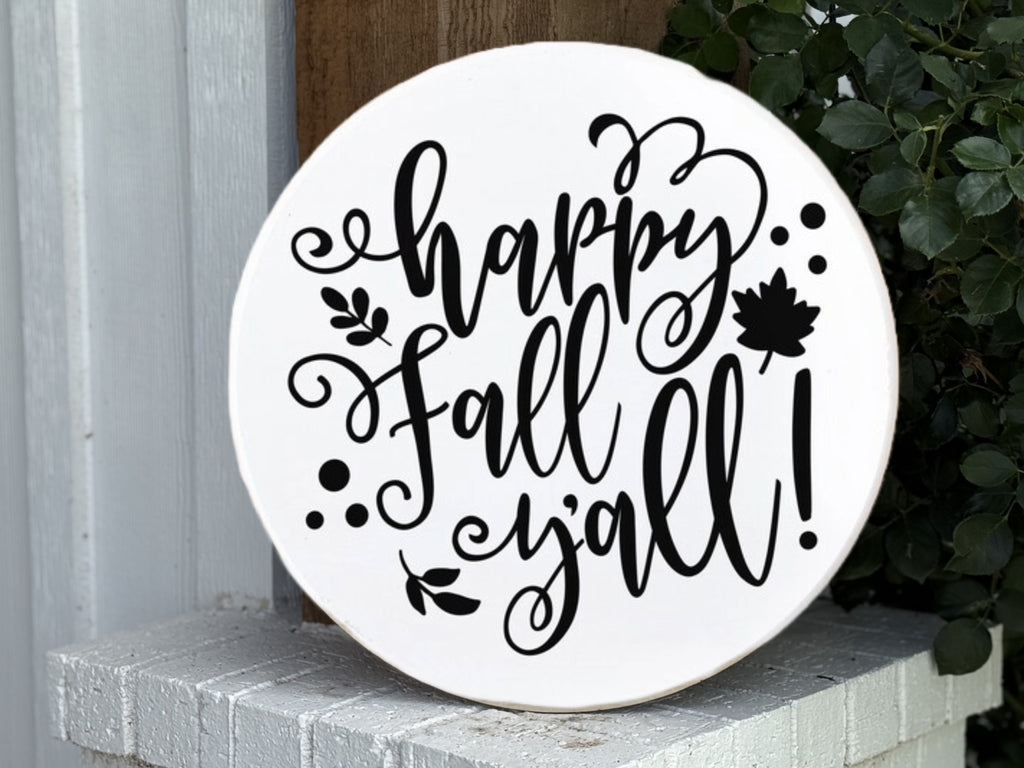 A round white sign with decorative black script that reads happy Fall yall! with leaf and swirl designs, propped on a white brick ledge next to green foliage.