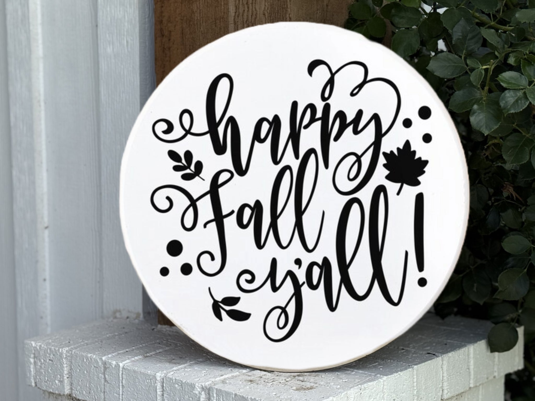 A round white sign with decorative black script that reads happy Fall yall! with leaf and swirl designs, propped on a white brick ledge next to green foliage.
