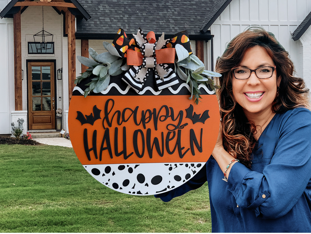 A round Halloween sign with happy HALLOWEEN in black letters, decorated with bats, black-and-white patterns, orange accents, faux greenery, and a large bow with candy corn print at the top.