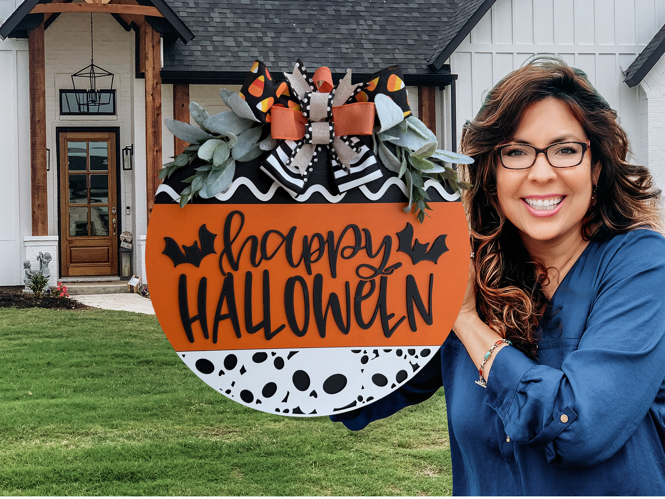 A round Halloween sign with happy HALLOWEEN in black letters, decorated with bats, black-and-white patterns, orange accents, faux greenery, and a large bow with candy corn print at the top.