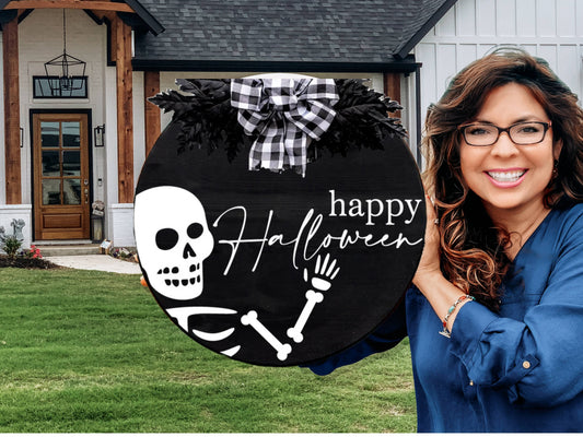 A woman smiles while holding a round black sign with a skeleton waving and the words “happy Halloween” in white script, standing outside a modern house with a green lawn.