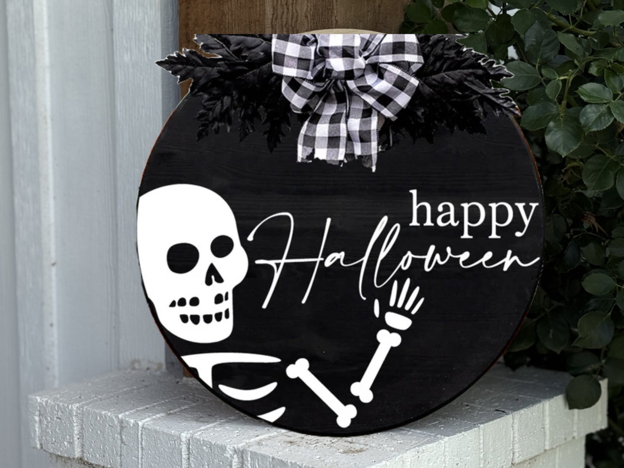 A round black sign with a white skeleton waving and the words happy Halloween in white script, decorated with black foliage and a black-and-white plaid bow at the top. The sign sits on a white brick surface.