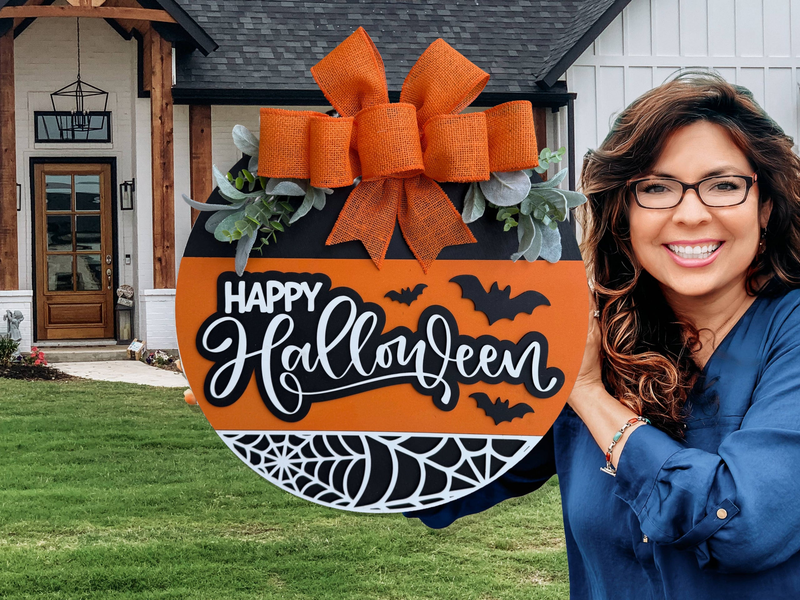 A smiling woman with long brown hair and glasses holds a round Happy Halloween sign decorated with bats, spiderwebs, greenery, and an orange bow, standing in front of a modern house with a green lawn.