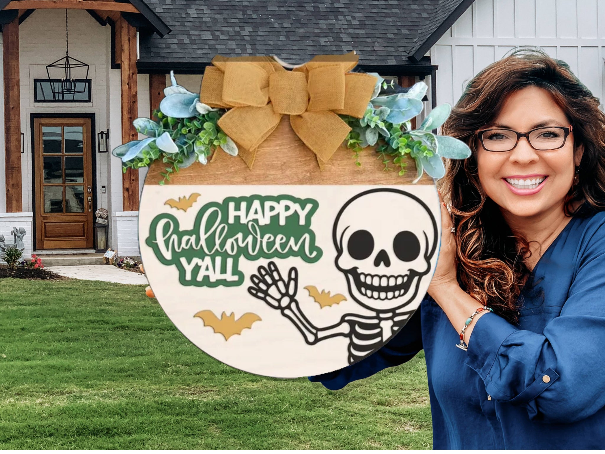A smiling woman with long brown hair and glasses holds a wooden sign decorated with a bow, greenery, a skeleton, bats, and the text Happy Halloween Yall in front of a house.