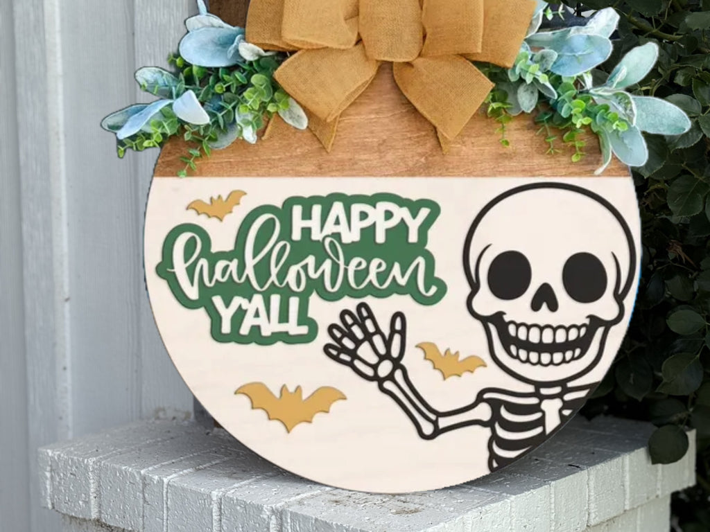 A round wooden sign decorated with greenery, a tan bow, and featuring a smiling skeleton, yellow bats, and the words Happy Halloween Yall in playful green and white letters.